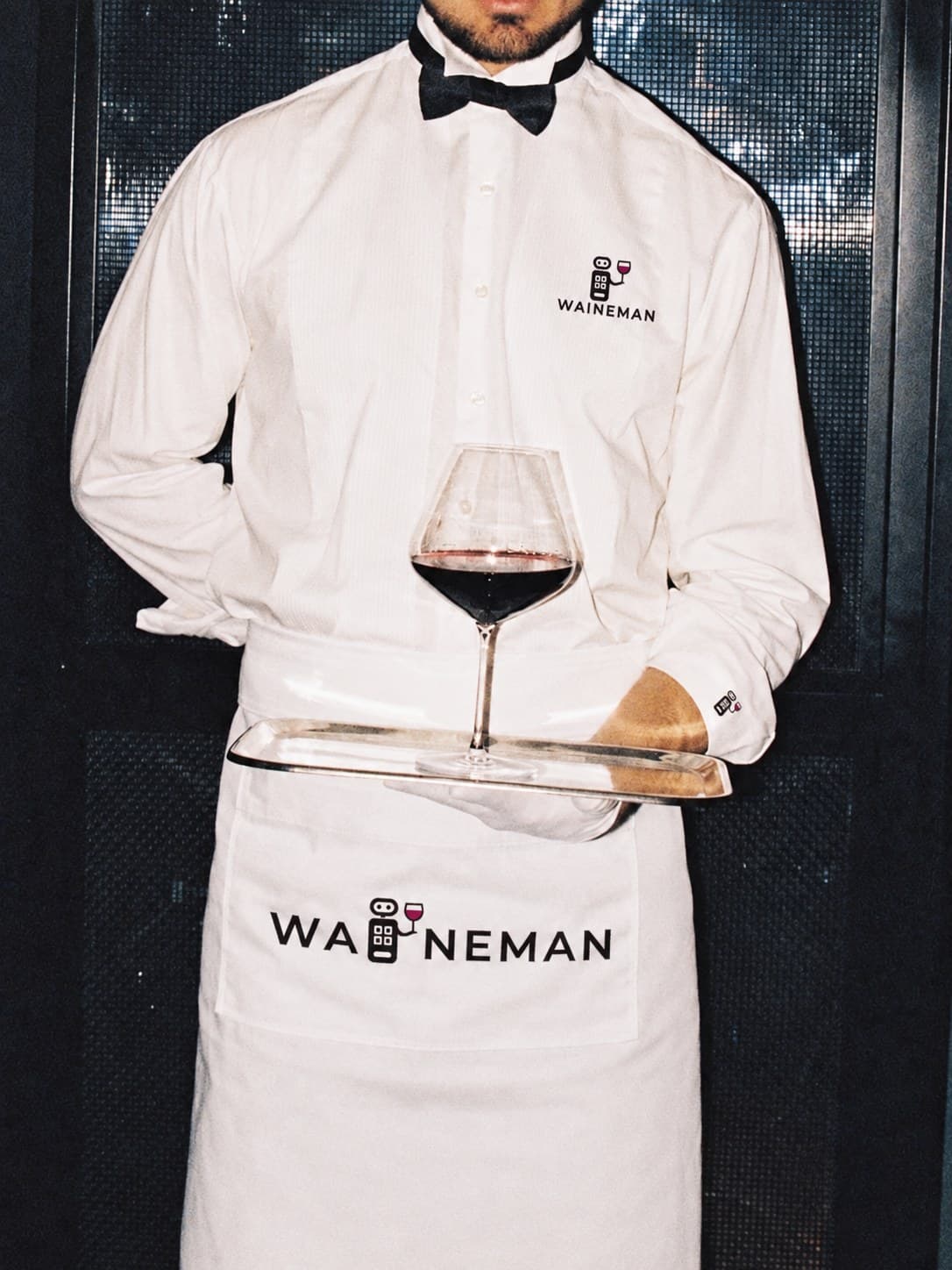Een ober in een wit overhemd en schort met het WAINEMAN-logo draagt een glas rode wijn op een dienblad
