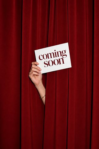 Een hand houdt een kaartje Coming soon door een rood gordijn